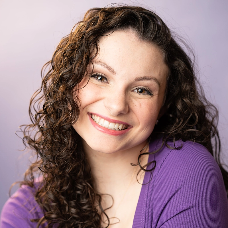Katie Krasovec, a young woman with long, dark, curly hair, smiles brightly at the camera. She is wearing a purple top, and the soft lavender background creates a warm and cheerful atmosphere.