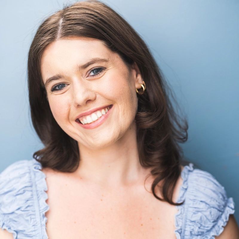 Katie Spiropoulos, a young woman with fair skin and wavy brown hair, smiles warmly. She is wearing a light blue top with ruffled edges and small gold hoop earrings. The background is a solid light blue, and the lighting is bright and even.