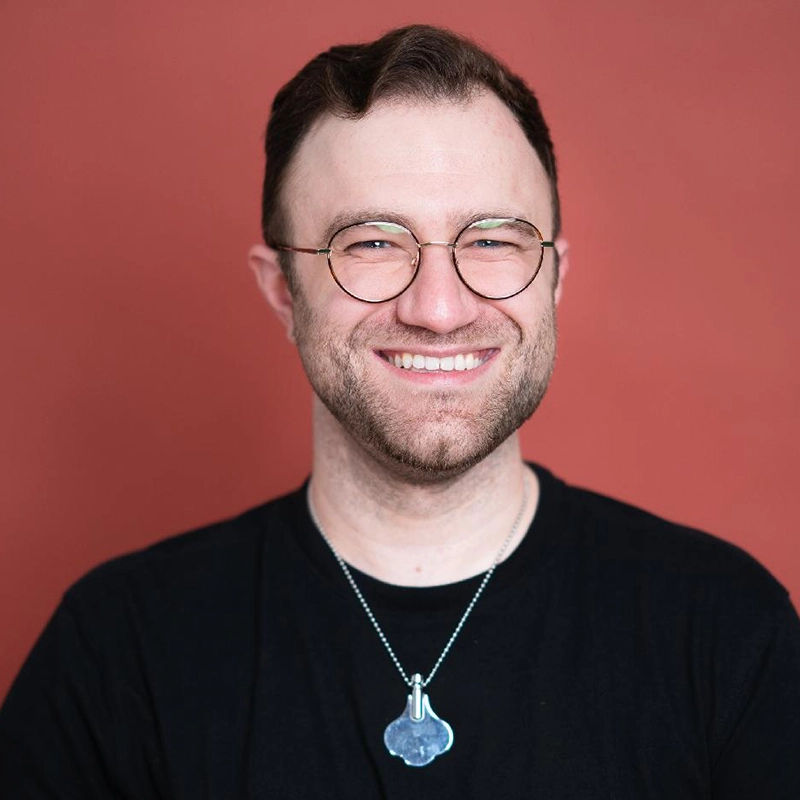 A smiling man with short brown hair, a trimmed beard, and round glasses stands against a reddish-brown background. He wears a black shirt and a silver pendant necklace by Francesca Pastore, featuring a unique, artistic shape.