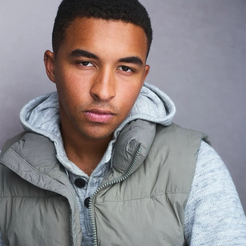 Steven Thomas, a young man with short hair, wears a light gray hoodie under an olive-green puffer vest. He looks directly at the camera with a serious expression against a simple gray background.