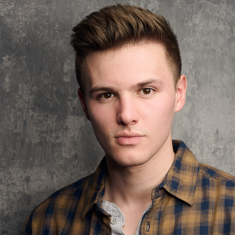 A young man with short, neatly styled brown hair stares seriously at the camera. He wears a brown and blue plaid shirt over a light gray T-shirt. The background is a textured, gray concrete wall, giving the image a professional, studio-like feel.