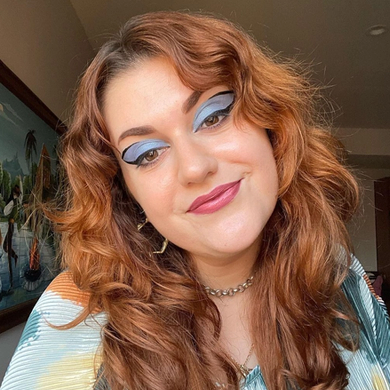 Abby Wolanin, with wavy auburn hair, smiles softly. She wears bold blue and black winged eyeshadow, pink lipstick, a silver necklace, and a striped top. The background reveals part of a painting with trees and water in a softly lit room.