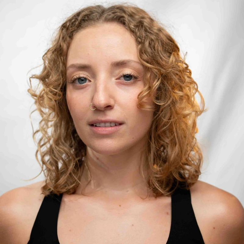 Melissa Tormene, a young woman with shoulder-length, curly blonde hair and blue eyes, stands against a plain white background. She wears a black tank top and has a small nose ring in her left nostril. Her expression is neutral and calm.