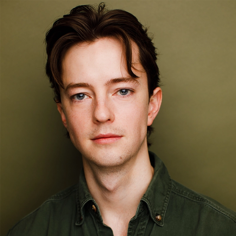 A young man with fair skin, blue eyes, and brown hair parted in the middle faces the camera with a neutral expression. Travis Flynt wears a green button-up shirt and stands against an olive-green background.