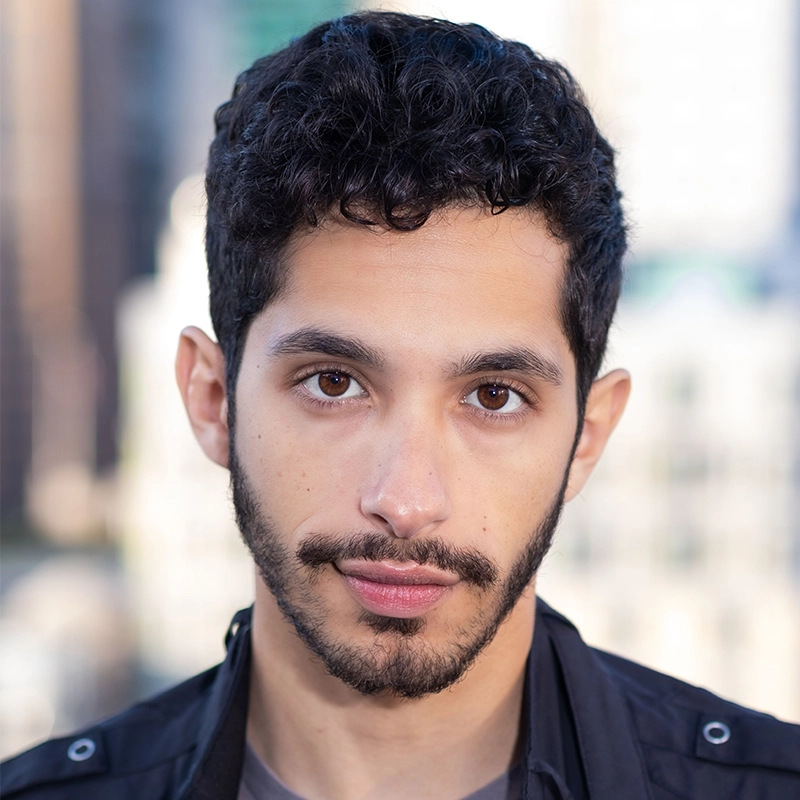 A young man with short curly black hair, light brown skin, and a trimmed beard looks directly at the camera. He wears a black jacket over a gray shirt. The blurred background, bright with sunlight and tall buildings, echoes Abby Wolanin’s urban energy.