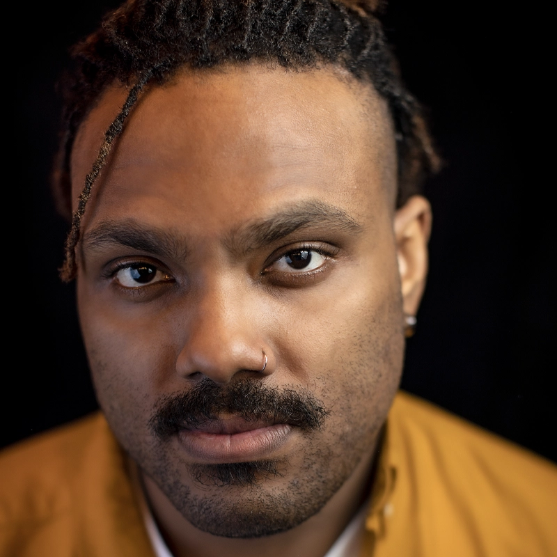 A man with medium brown skin, a mustache, short beard, and a nose ring looks directly at the camera. Sporting Chris Tucker–inspired short dreadlocks, he wears a mustard yellow shirt with a black background behind him.
