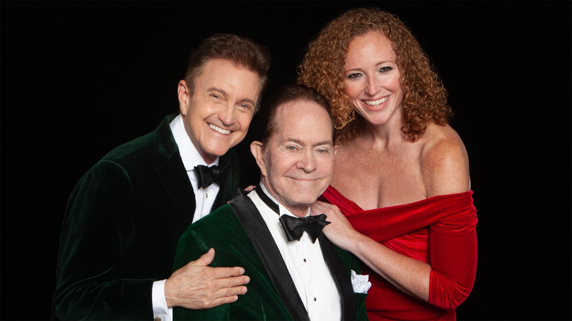 Three adults pose closely against a black background. At an Annual Concert honoring Alec Wilder’s music, two men in green velvet tuxedos and bow ties stand left and center, while a woman with curly red hair in a red off-shoulder dress smiles warmly on the right.