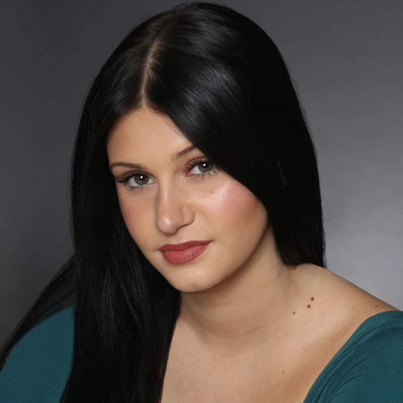 Lorelei Vega, a young woman with long, straight black hair and green eyes, gazes at the camera with a neutral expression. She has light skin, subtle makeup, and wears a teal top against a dark gray background.