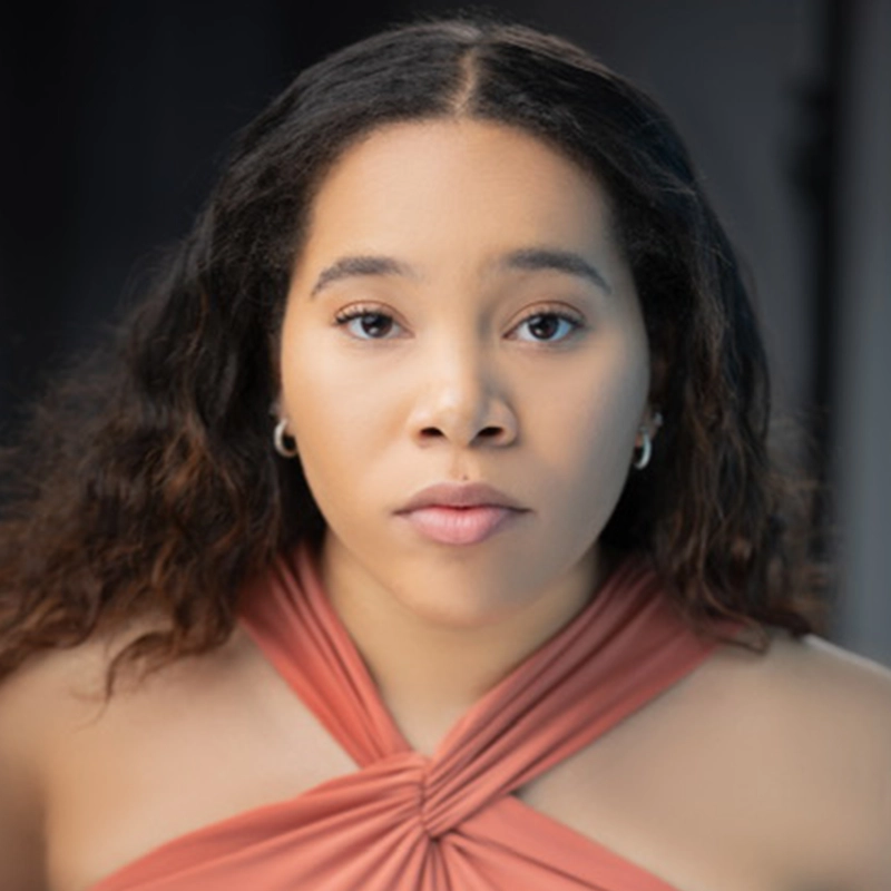 Vivica Powell, a young woman with medium skin tone and long, wavy brown hair, stares directly at the camera. She wears a peach-colored halter top and small hoop earrings, her calm expression set against a softly blurred, dark background.