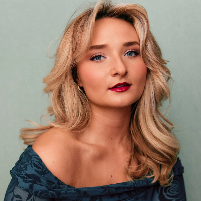 A young woman, Morgan Vybihal, with wavy blonde hair and blue eyes looks into the camera. She wears bold red lipstick and a blue off-the-shoulder floral top. The background is a soft, muted green with flattering, gentle lighting.