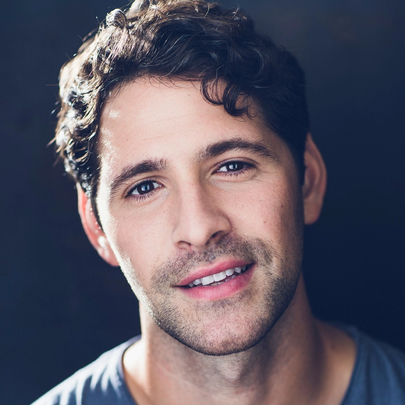 Dylan Wallach, a man with wavy dark hair and light skin, smiles softly at the camera. He has stubble, brown eyes, and is wearing a grey t-shirt. The dark, blurred background highlights his face with gentle lighting.