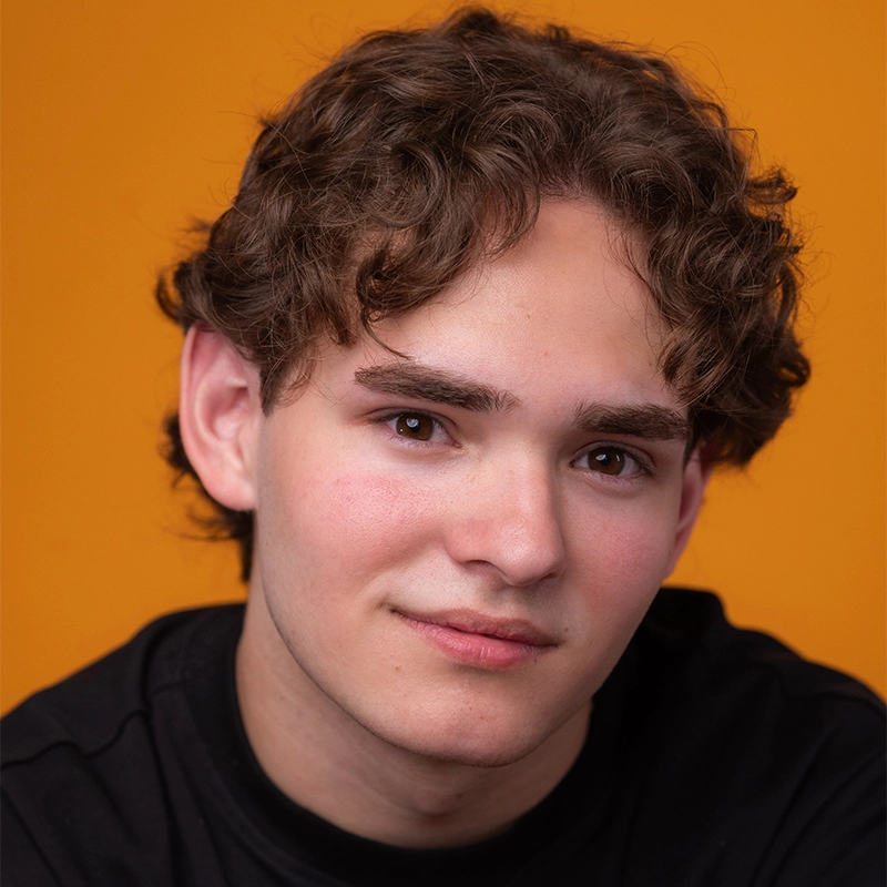 A young person with wavy brown hair and fair skin looks at the camera, smiling gently. Wearing a black shirt, Katie Krasovec is posed against an orange background in this close-up portrait focusing on the face and shoulders.
