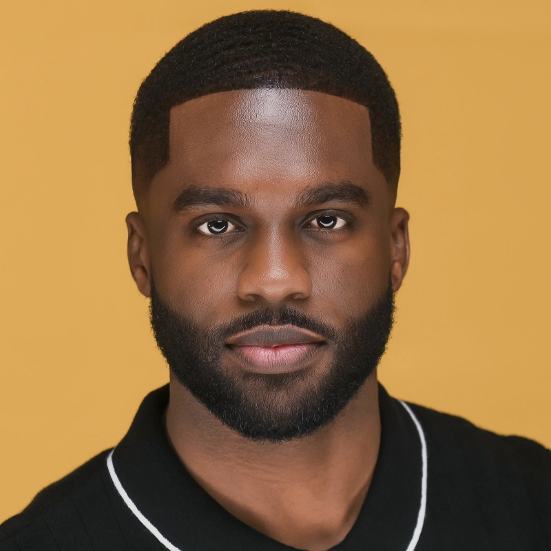 Adarian Williams, with short, closely-cropped hair and a trimmed beard, looks directly at the camera. He wears a black collared shirt with white piping against a solid yellow background, his expression neutral and confident.