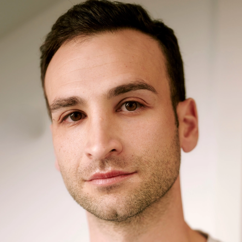 A man with short dark hair and trimmed facial stubble looks directly at the camera with a neutral expression. He has brown eyes and fair skin. The softly blurred background brings the focus to his face, highlighting Harley Yanoff’s features.