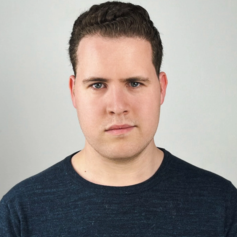 Tal Marcello Zamir, a young man with short, dark, wavy hair and fair skin, stares seriously at the camera. He wears a dark blue crewneck sweater against a plain, light gray background.