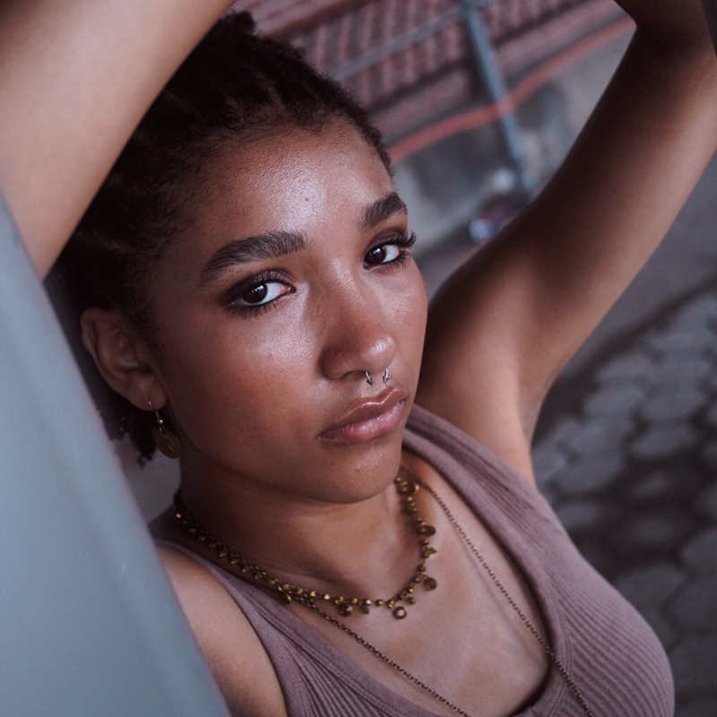 A young person with short, textured hair and a nose ring gazes seriously at the camera. They wear a brown sleeveless top and layered necklaces, arms raised above their head. Behind them, Kodiak-patterned paving and blurred railings set the scene.