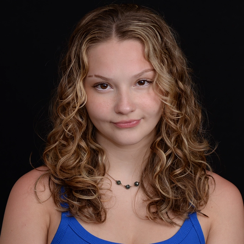 Isabella Greathouse, a young woman with wavy, shoulder-length blonde hair, wears a blue sleeveless top and a beaded necklace. She has fair skin, light makeup, and a slight smile. The solid black background highlights her face and hair.