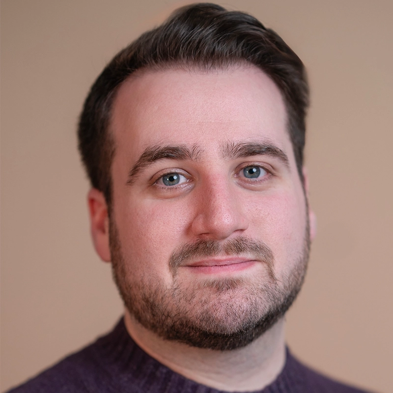 A man with fair skin, short brown hair, and a trimmed beard is facing the camera and smiling slightly. Mickey Wirtz is wearing a dark purple sweater against a plain beige background, creating a professional headshot style.