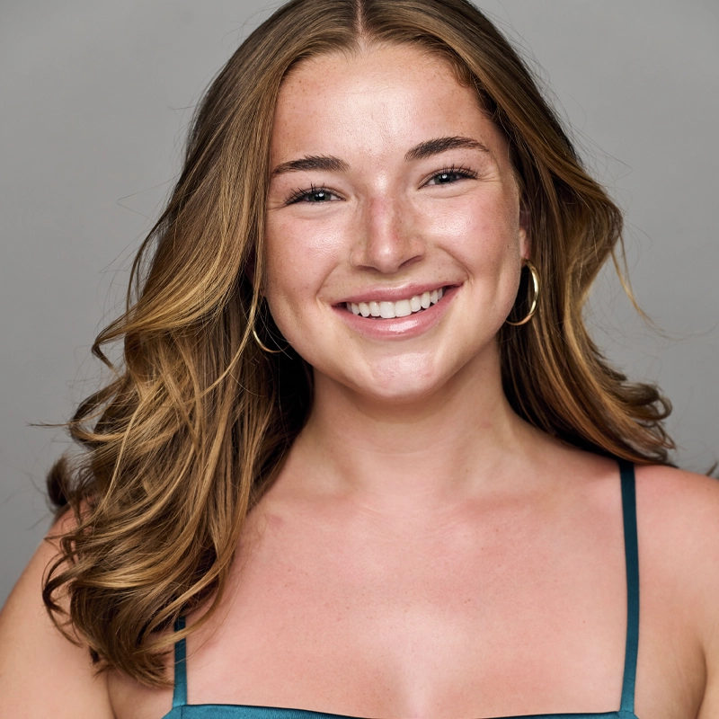 A young woman with wavy, light brown hair, wearing hoop earrings and a teal strappy top, smiles warmly at the camera against a plain gray background. Her skin is lightly freckled and she appears relaxed and happy.