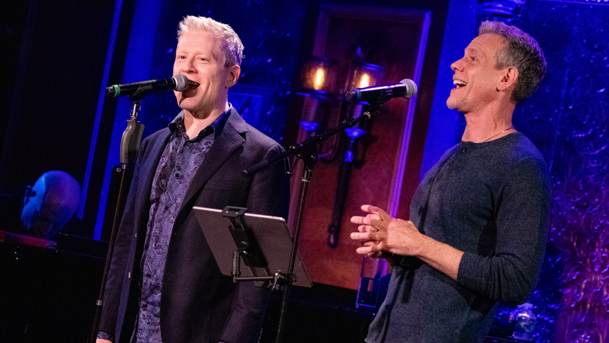 Anthony Rapp and Adam Pascal stand side by side on stage, each singing into a microphone. Both appear enthusiastic amid blue and purple lighting and ornate decor, perfectly capturing the energy of their 
