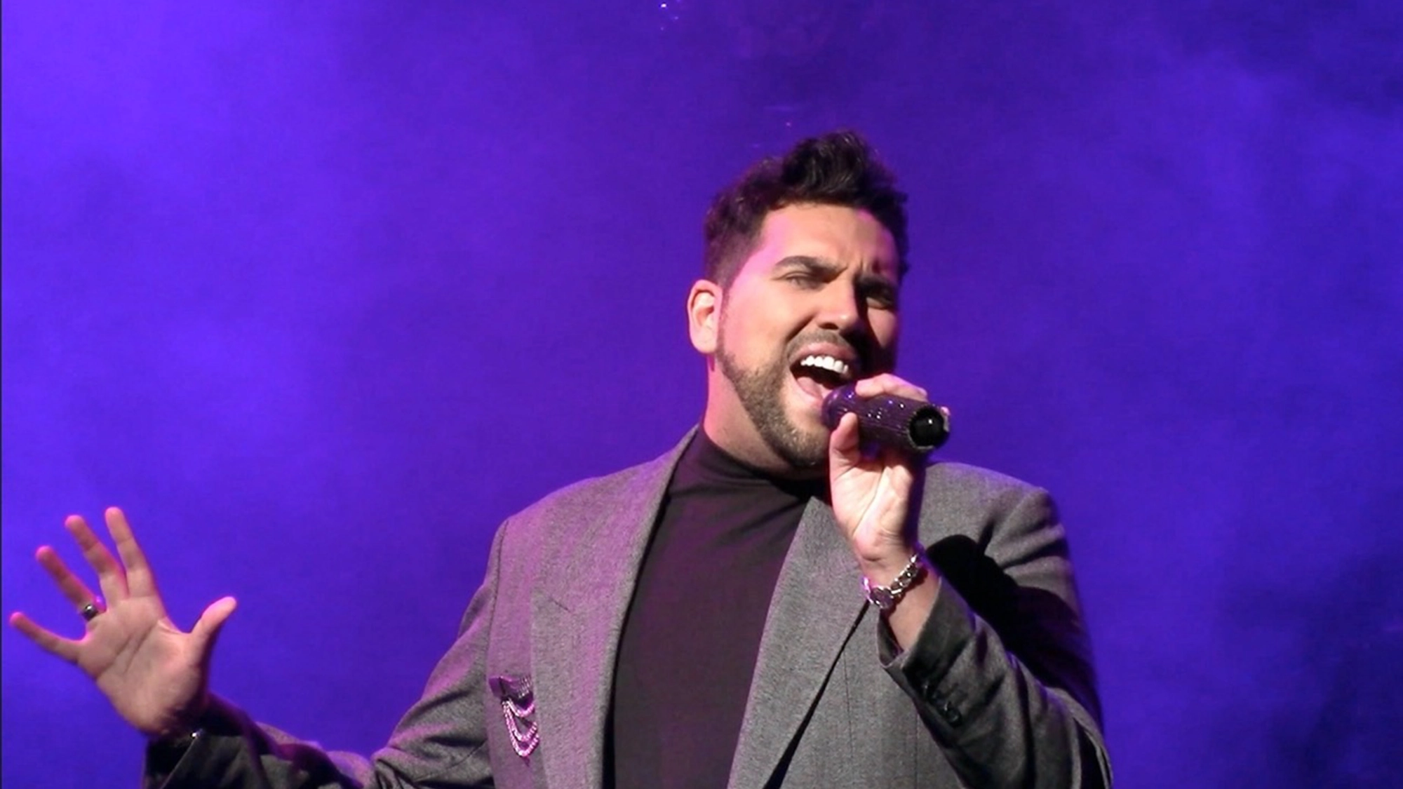 Adam Strong, dressed in a gray blazer over a black turtleneck, passionately sings into a microphone on stage. With dramatic gestures under purple Lights Up stage lighting and mist, he commands attention at this live performance.