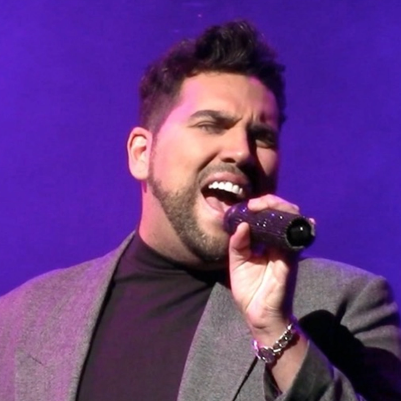 A man with short dark hair and a trimmed beard sings passionately into a microphone. He is wearing a gray blazer over a black shirt against a purple-lit background. His mouth is open wide, and his eyes are focused, showing emotion as he performs.