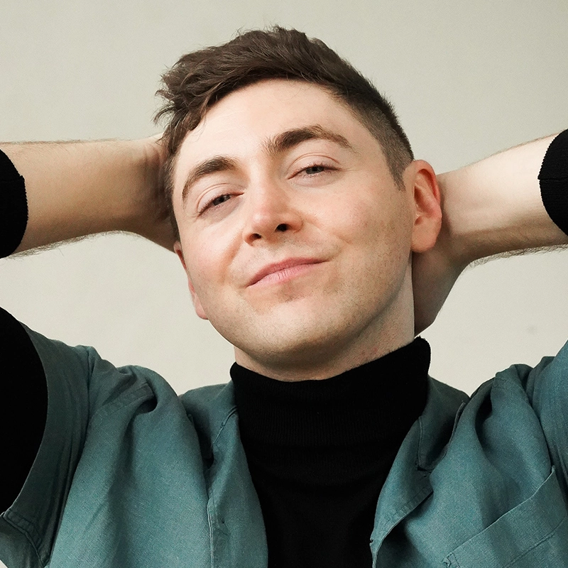 A person with short brown hair smiles softly, leaning back with both hands resting behind their head. They wear a green jacket over a black turtleneck. The background is plain and light-colored, creating a relaxed, casual atmosphere.