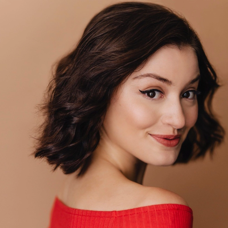 Sophie Aknin, a young woman with short, wavy brown hair, smiles over her shoulder. She has winged eyeliner, light makeup, and wears an off-the-shoulder red top against a plain beige background that keeps her as the clear focus.