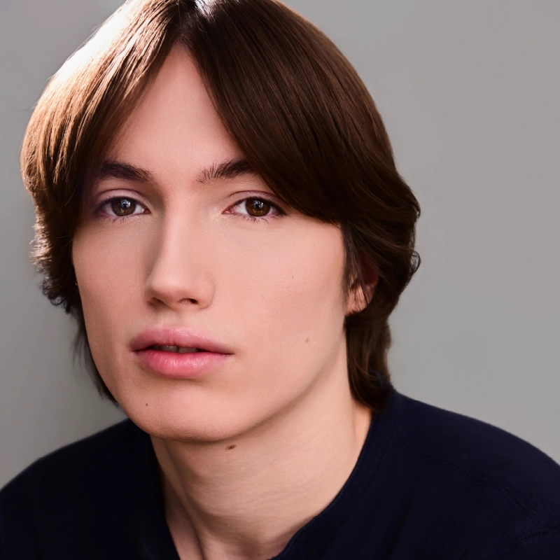 Rynn Stephenson, a young person with fair skin and straight brown hair parted in the middle, looks at the camera. They have brown eyes, smooth skin, and wear a dark shirt against a plain gray background that highlights their face.