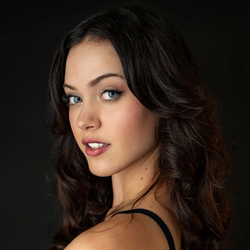 A young woman with fair skin, long wavy dark brown hair, and striking blue eyes looks over her shoulder toward the camera. She wears natural makeup and a black tank top, captured in classic RJ Vandenbrouck style against a plain dark background.