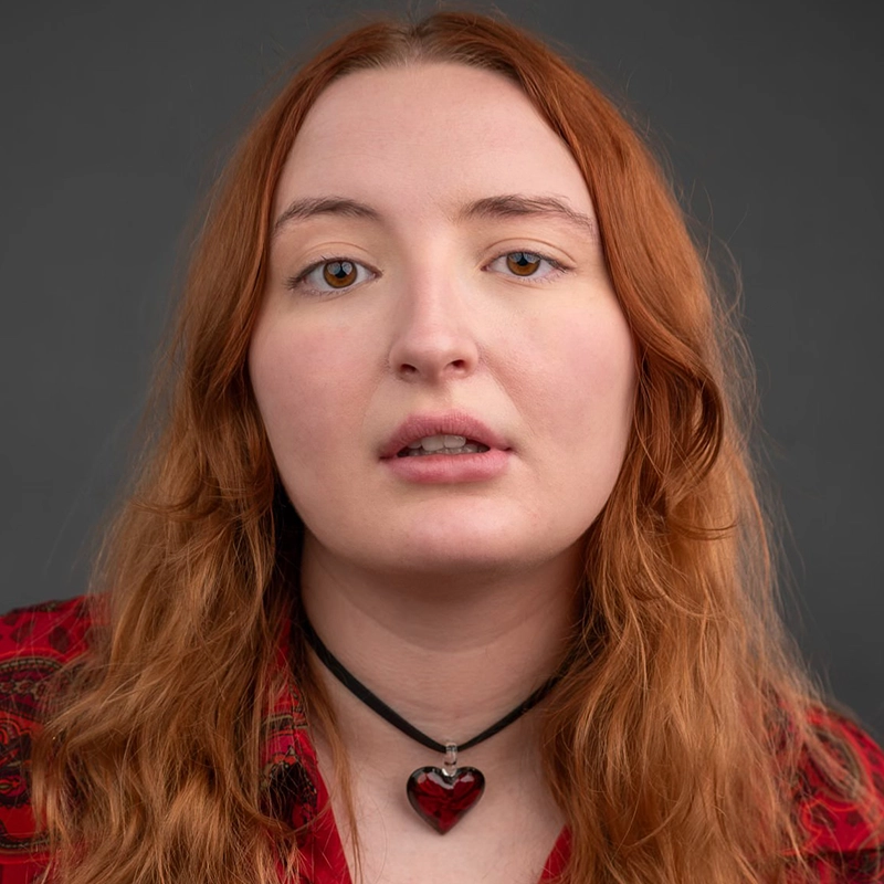 Rynn Stephenson, with long, wavy reddish-brown hair, wears a red patterned shirt and a black choker with a red heart pendant. Fair-skinned and neutral-faced, they look directly at the camera against a plain dark gray background.