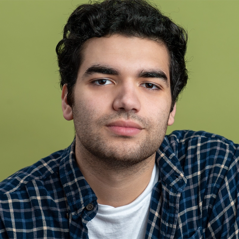 A young man with short, curly dark hair and a trimmed beard looks at the camera with a neutral expression. Justin Amaro is wearing a blue plaid shirt over a white t-shirt, set against a solid light green background.