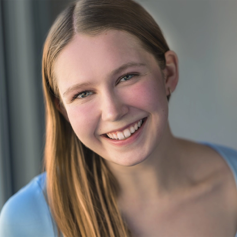 A young woman with straight, light brown hair and fair skin smiles warmly at the camera. She is wearing a light blue top and is positioned in soft natural light, with a blurred gray background. Her head is slightly tilted, and her expression is joyful.