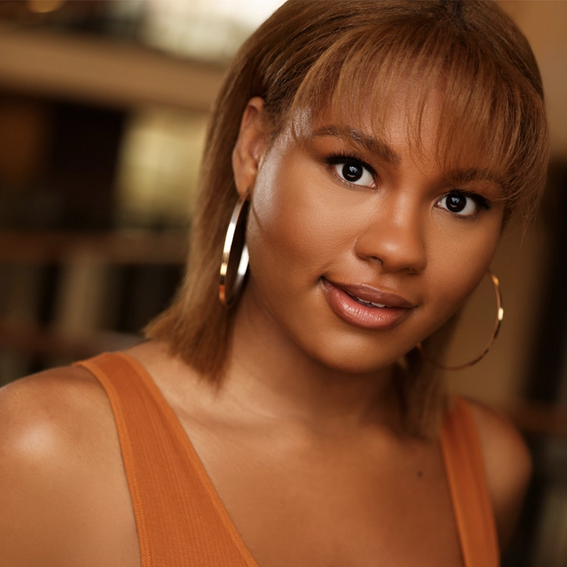 Anyae Anasia, a young woman with straight, light brown hair and bangs, smiles softly. She wears large gold hoop earrings and an orange sleeveless top. The background is softly blurred in warm tones, drawing focus to her face.