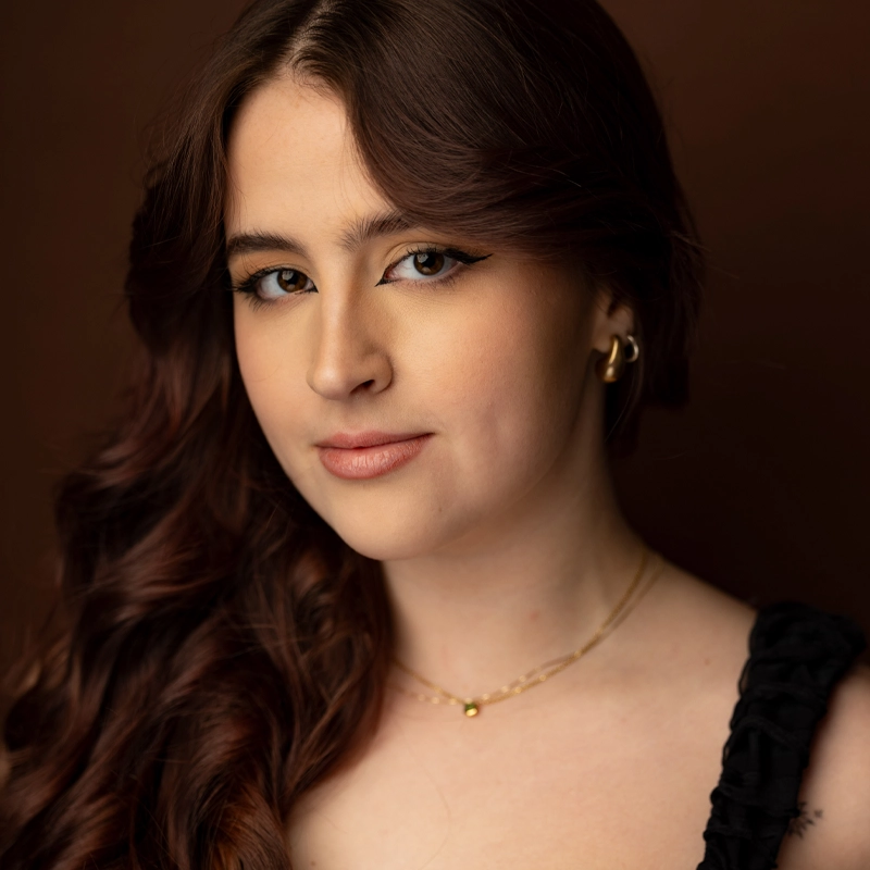 A young woman with long, wavy brown hair poses against a brown background. She wears a black top, gold hoop earrings, and two delicate gold necklaces. Her makeup is natural with winged eyeliner, and she has a slight, confident smile.