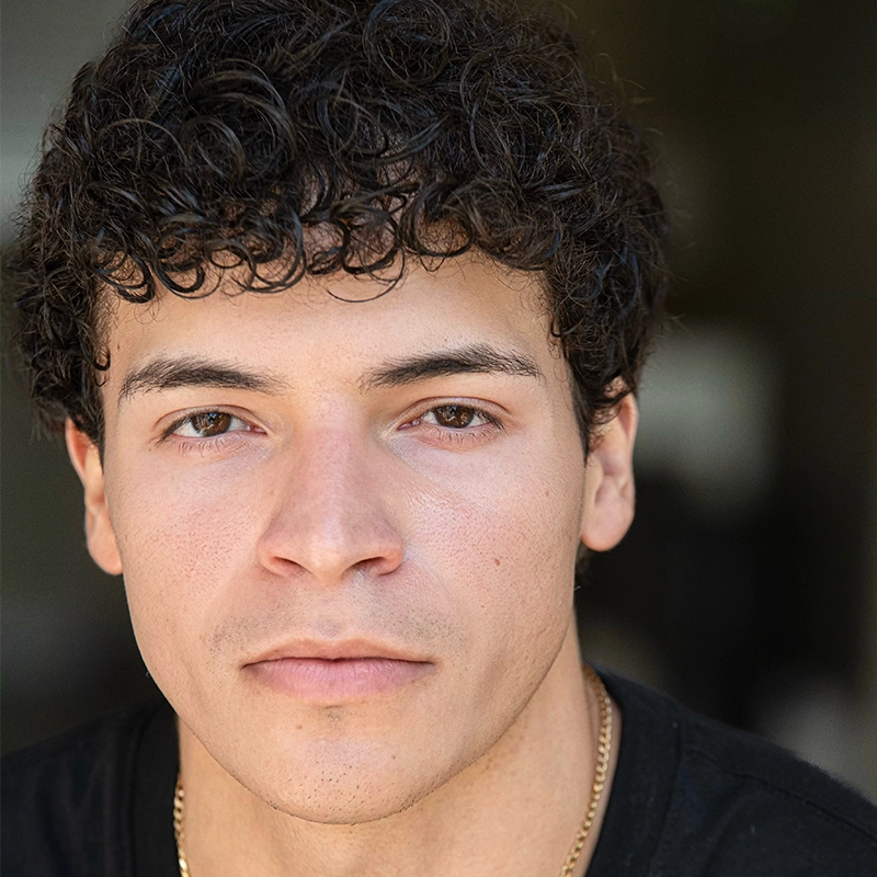 Anthony Williams, a young man with short, curly dark hair and light brown skin, looks directly at the camera with a neutral expression. He wears a black shirt and a thin gold chain necklace. The blurred, dark background draws focus to his face.
