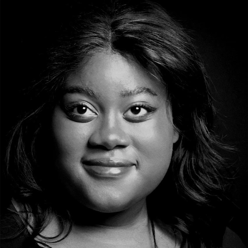 A black and white portrait of Kenzie Woolridge with wavy, shoulder-length hair. She is looking slightly to the side and smiling softly. The lighting highlights her facial features against a dark background.