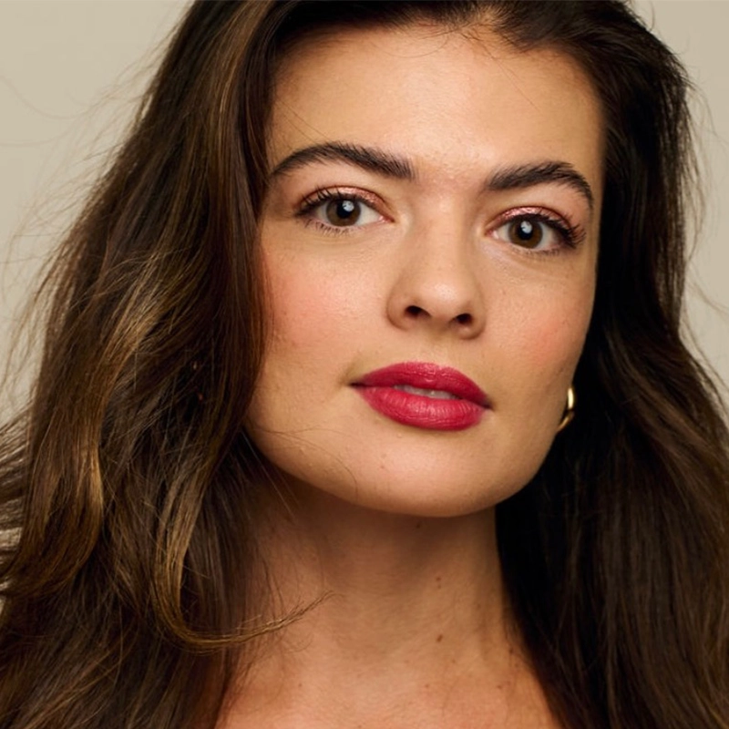 Hannah Thompson, with long wavy brown hair and fair skin, looks directly at the camera. She has full eyebrows, brown eyes, pink lipstick, and wears a gold hoop earring against a neutral background.