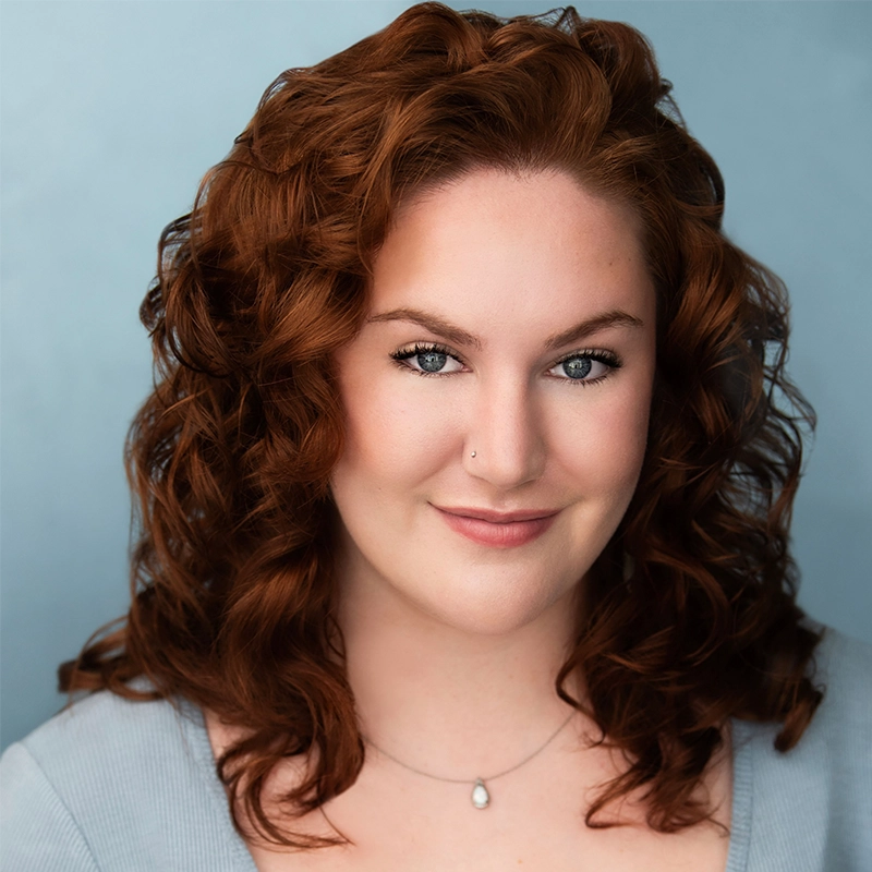 Sahara Williams, a woman with fair skin and curly red hair, smiles softly at the camera. She wears a light gray top, a delicate teardrop pendant necklace, and has a small nose piercing against a soft, pale blue background.