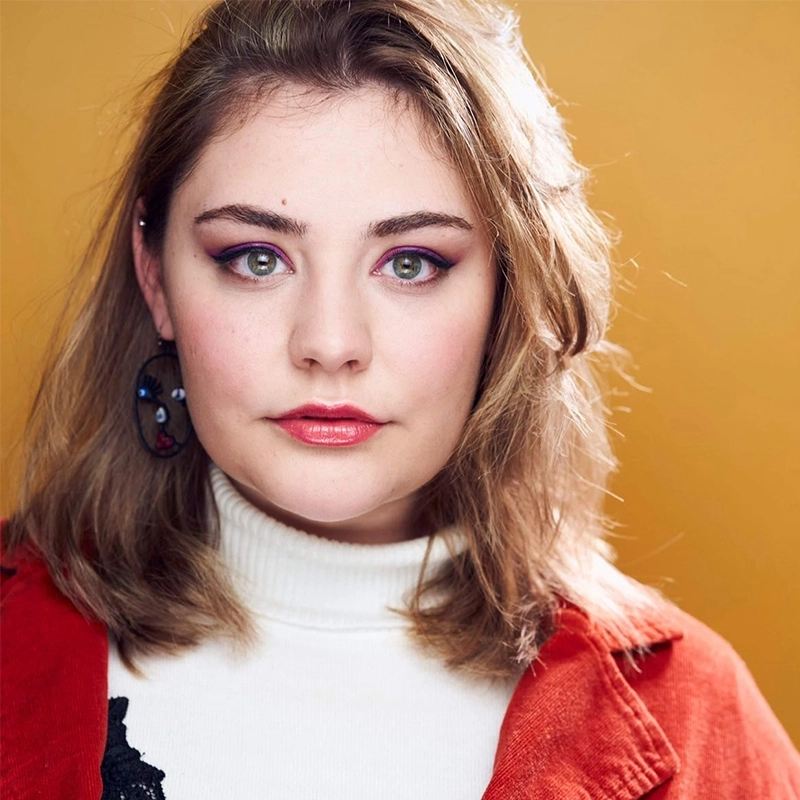 Caroline Weston, a young woman with light skin and straight, light brown hair, looks directly at the camera. She wears a white turtleneck, red jacket, bold eye makeup, and a single dangling earring against a mustard yellow background.