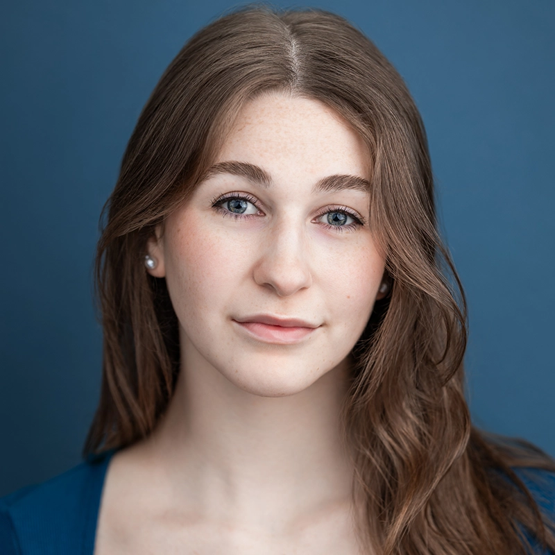 Caroline Weston, a young woman with fair skin, long brown hair, and blue eyes, looks directly at the camera. She wears a blue top and small pearl earrings against a solid blue background with soft, even lighting highlighting her features.