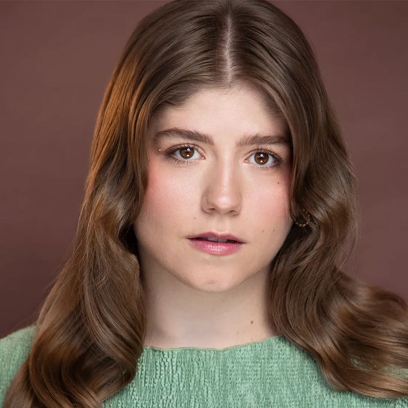 A young woman with long, wavy brown hair wearing a light green textured top looks directly at the camera. She has fair skin, natural makeup, and a neutral expression. The background is a muted brown color.