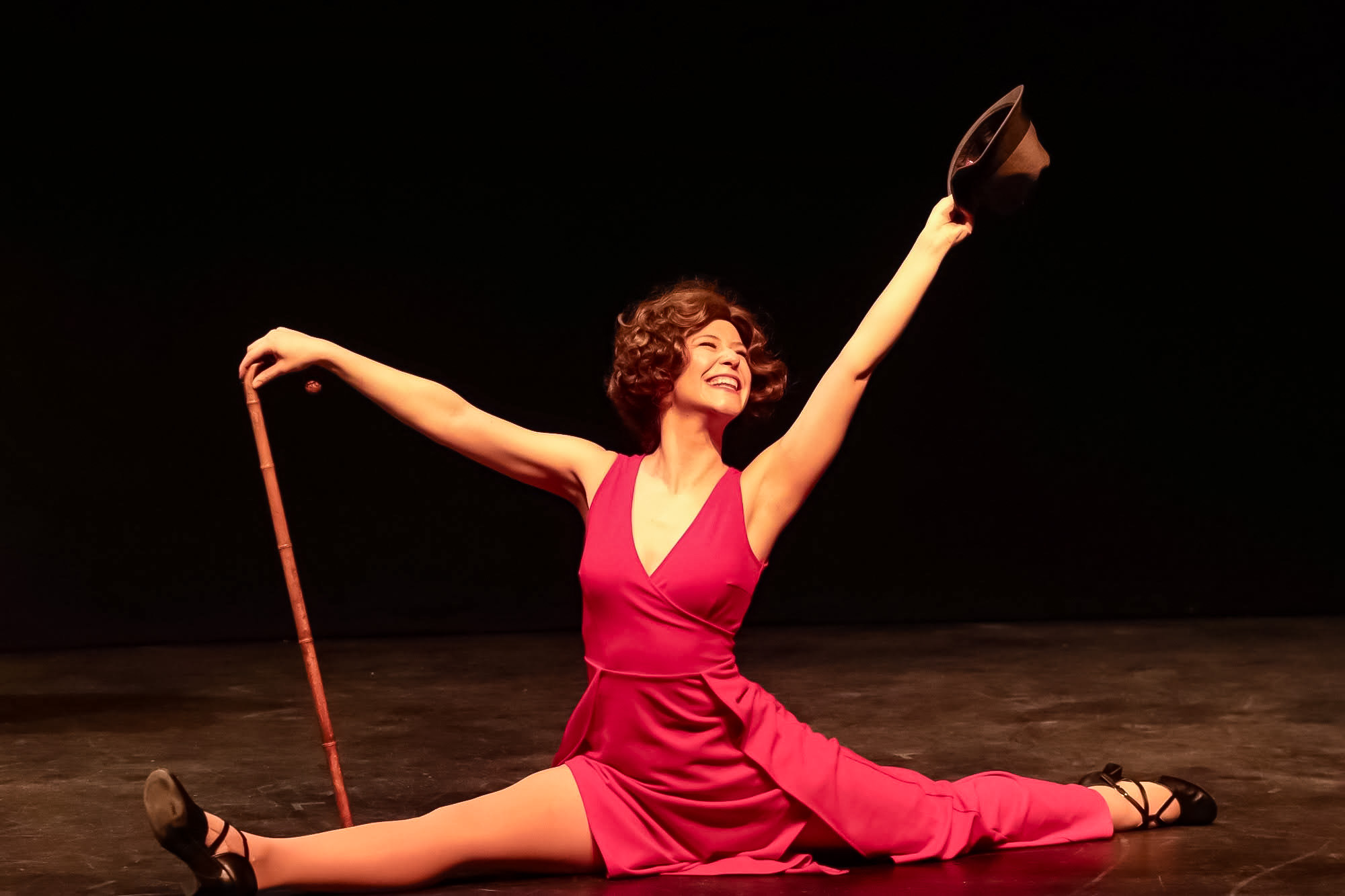 A woman in a sleeveless red dress does the splits on stage, holding a cane and raising a hat. She smiles brightly, her short curly hair shining in the spotlight. This vibrant scene is from the new musical Corinne’s Heart by Charles Mandracchia.