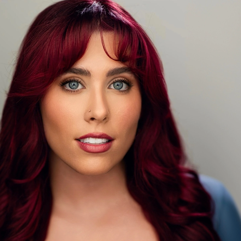 A person with long, wavy burgundy hair and blue eyes faces the camera. They have natural makeup, groomed eyebrows, and wear a subtle nose stud. The background is softly blurred in neutral tones, drawing focus to their face.