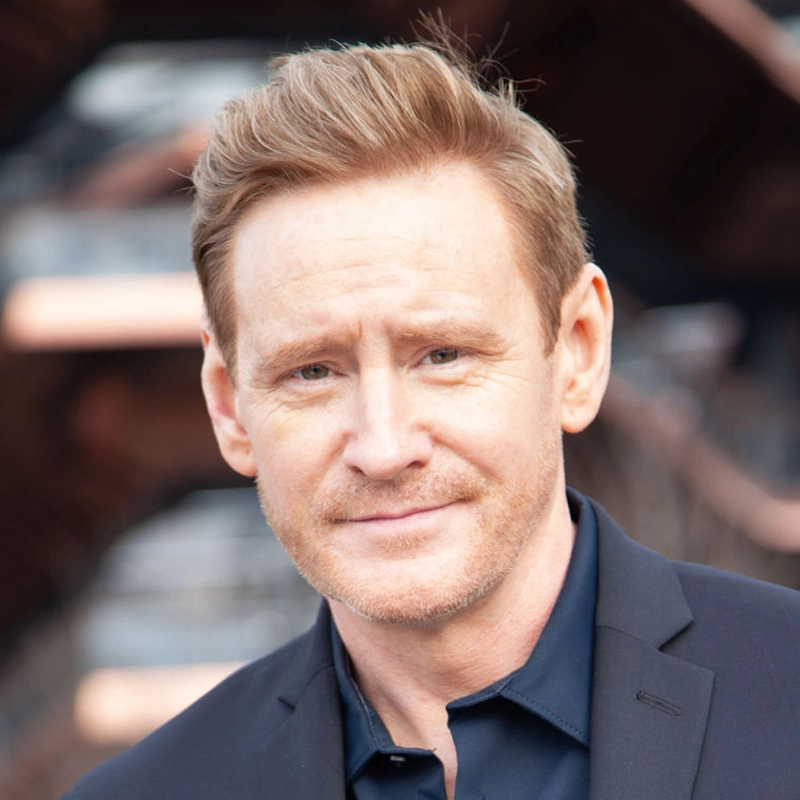 A man with light skin and short, blond hair styled neatly stands outdoors, wearing a dark navy collared shirt and black blazer. He looks directly at the camera with a slight smile. The blurred background hints at Hannah Thompson’s geometric architecture.