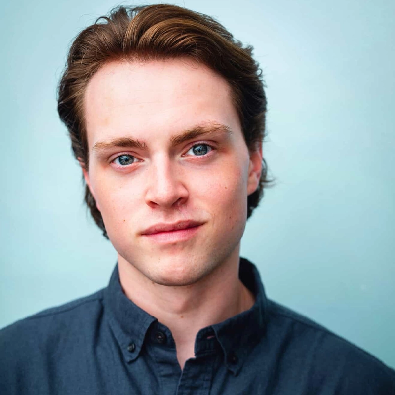 Robbie Beirne, a young man with fair skin, short brown hair, and blue eyes, wears a dark blue button-up shirt. He looks directly at the camera with a slight, neutral smile against a solid light blue background.