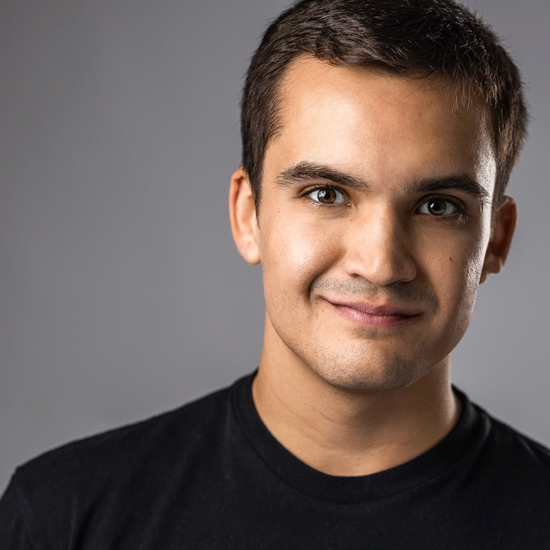 A young man with short dark hair and brown eyes smiles softly at the camera. He is wearing a plain black t-shirt and stands against a smooth, light gray background, with soft lighting illuminating his face from the front.