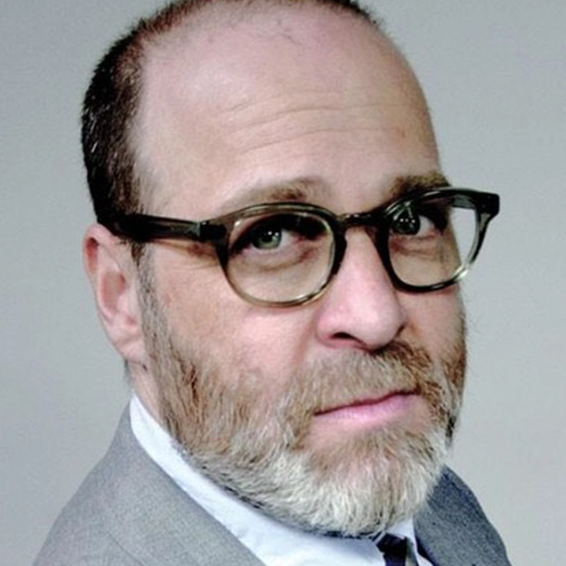 A middle-aged man with a receding hairline, gray beard, and mustache, resembling H. Jon Benjamin, wears dark, thick-rimmed glasses and a gray suit. He looks slightly past the camera with a neutral expression against a plain, light background.