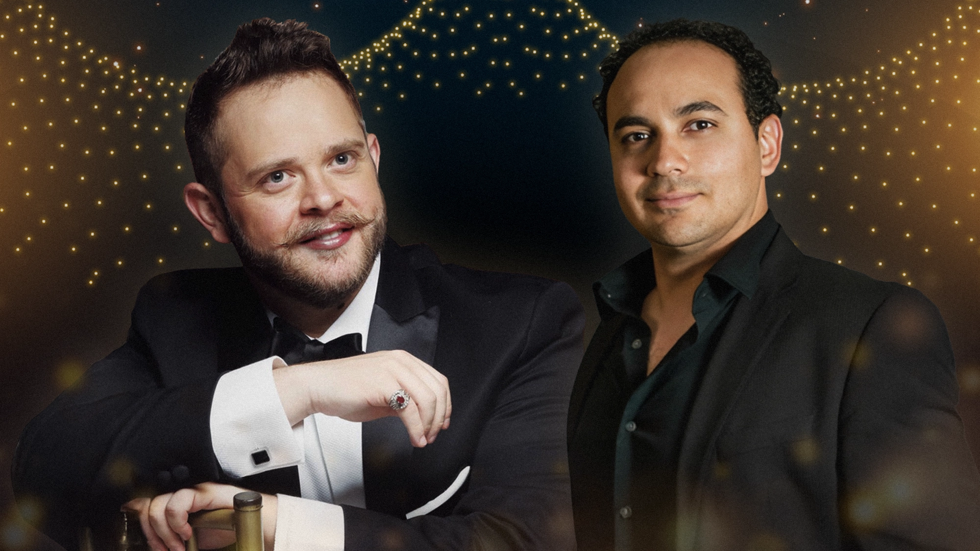 Two men in formal attire pose before golden string lights. Rodrigo de la Cadena, in a tuxedo, smiles with his hand on a chair, while José Adán Pérez stands beside him—both stars of Boleros de Medianoche—looking confidently at the camera.