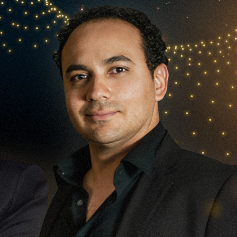 A man with short, curly black hair and medium skin tone wears a black suit jacket over a black shirt. He faces forward, slightly smiling, with soft golden lights and a dark background behind him, creating an elegant, formal atmosphere.